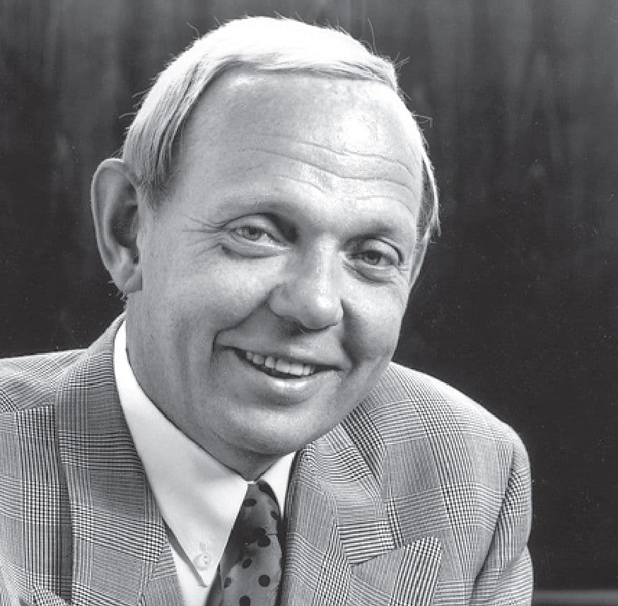 Black and white image of a smiling man in a plaid suit and polka-dot tie.