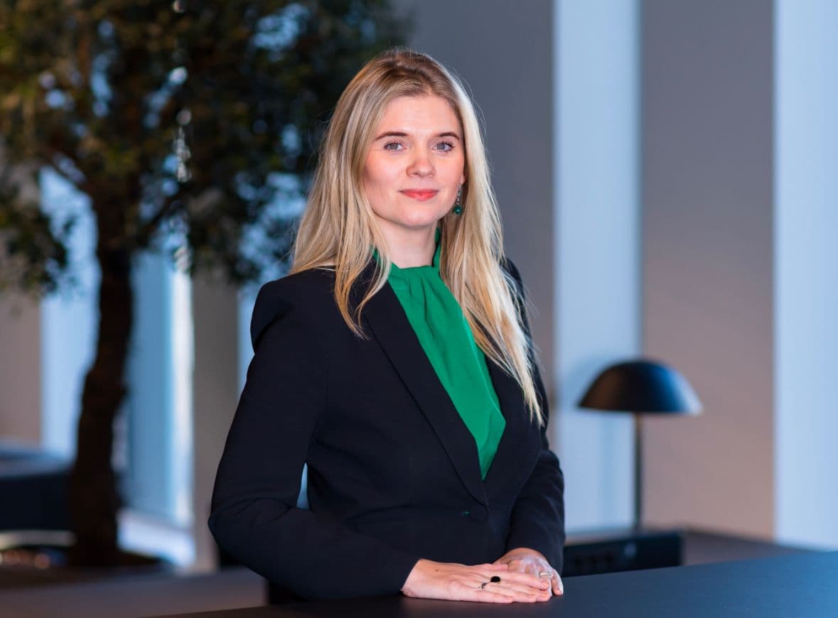 A woman with blonde hair wearing a black blazer and green top, leaning on a desk with a slight smile.