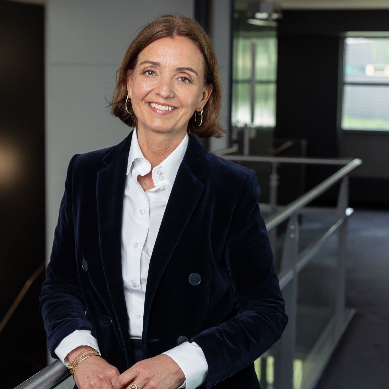 A smiling woman in a dark blue velvet blazer and white shirt leans on a railing.