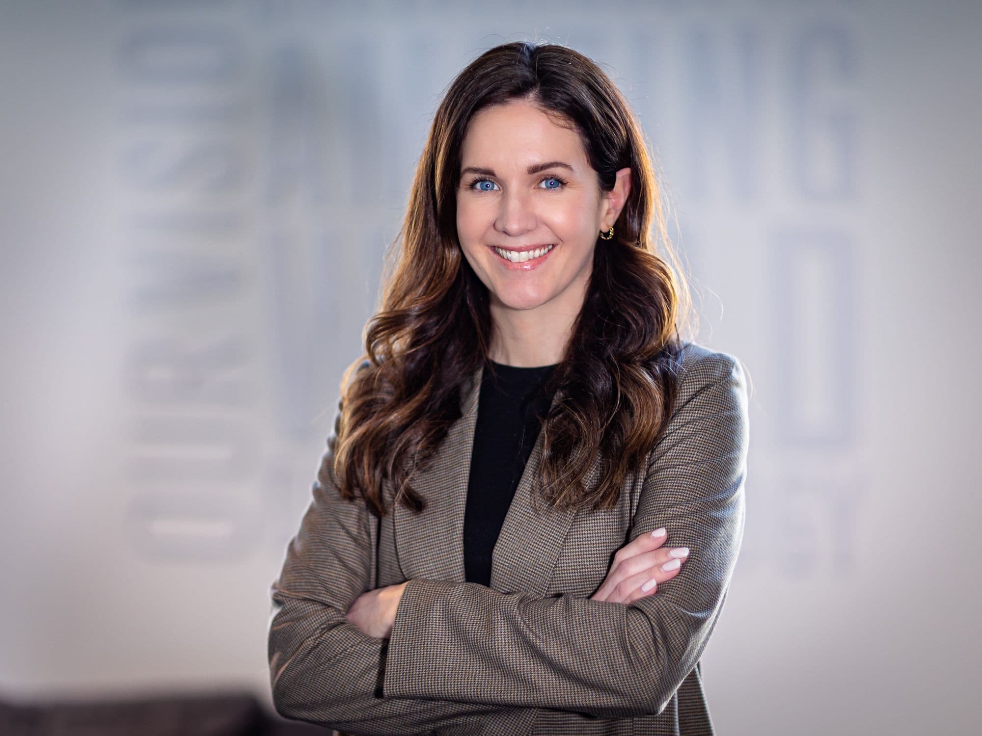 A smiling woman with brown wavy hair and blue eyes, wearing a brown blazer, stands with crossed arms.
