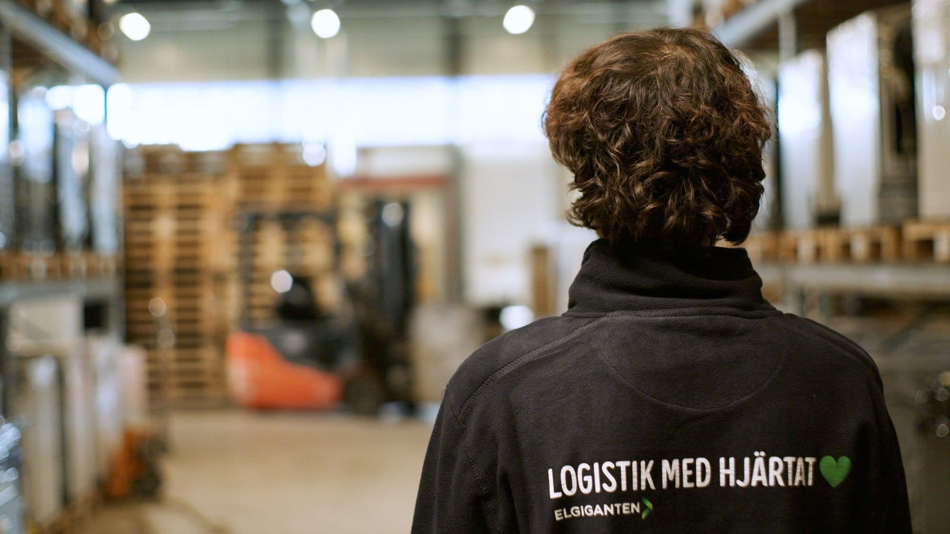 A person seen from behind in a warehouse, wearing a black jacket with "LOGISTIK MED HJÄRTAT" and a green heart printed on it.