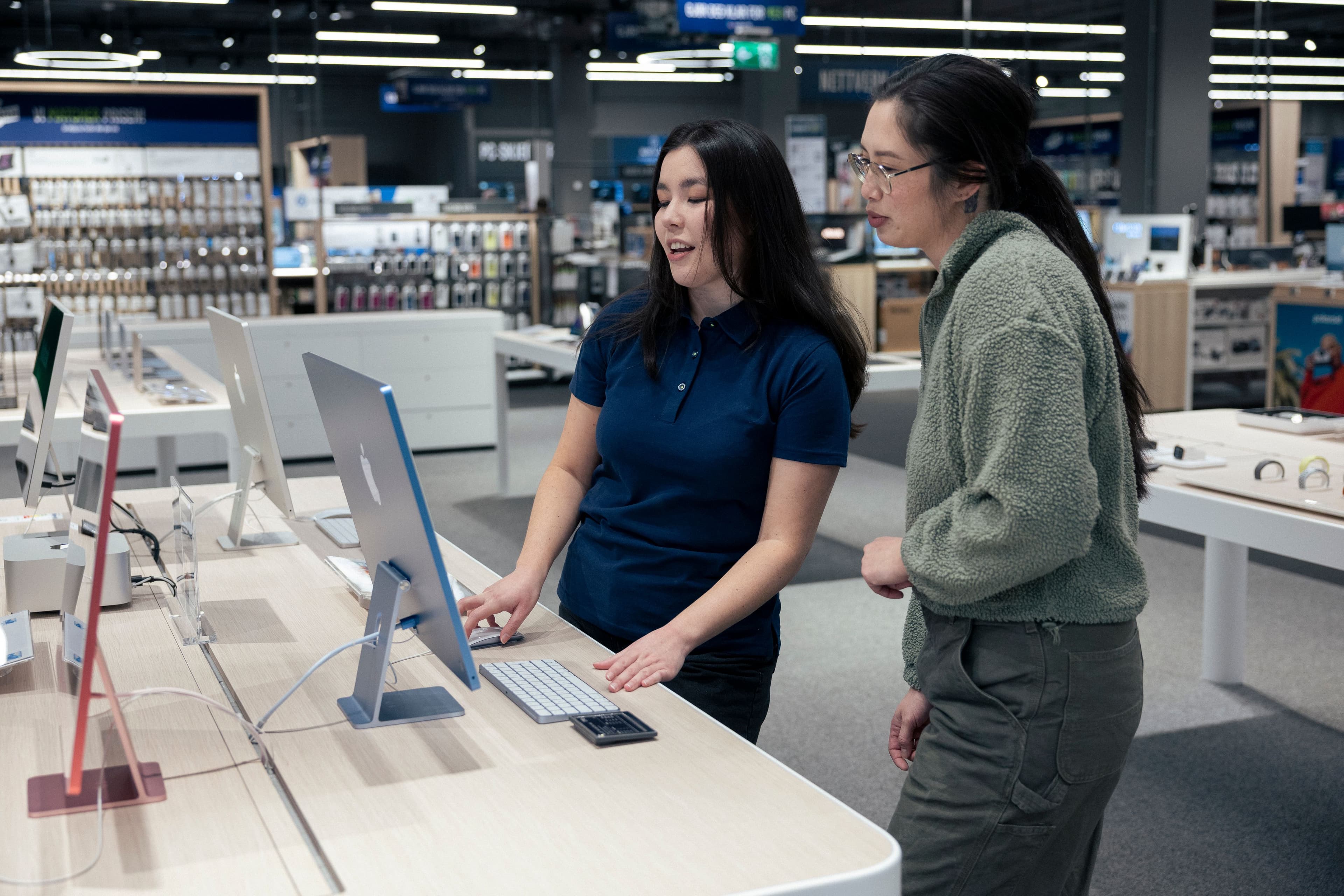 Two women examine a blue desktop computer on a display table in an electronics store.