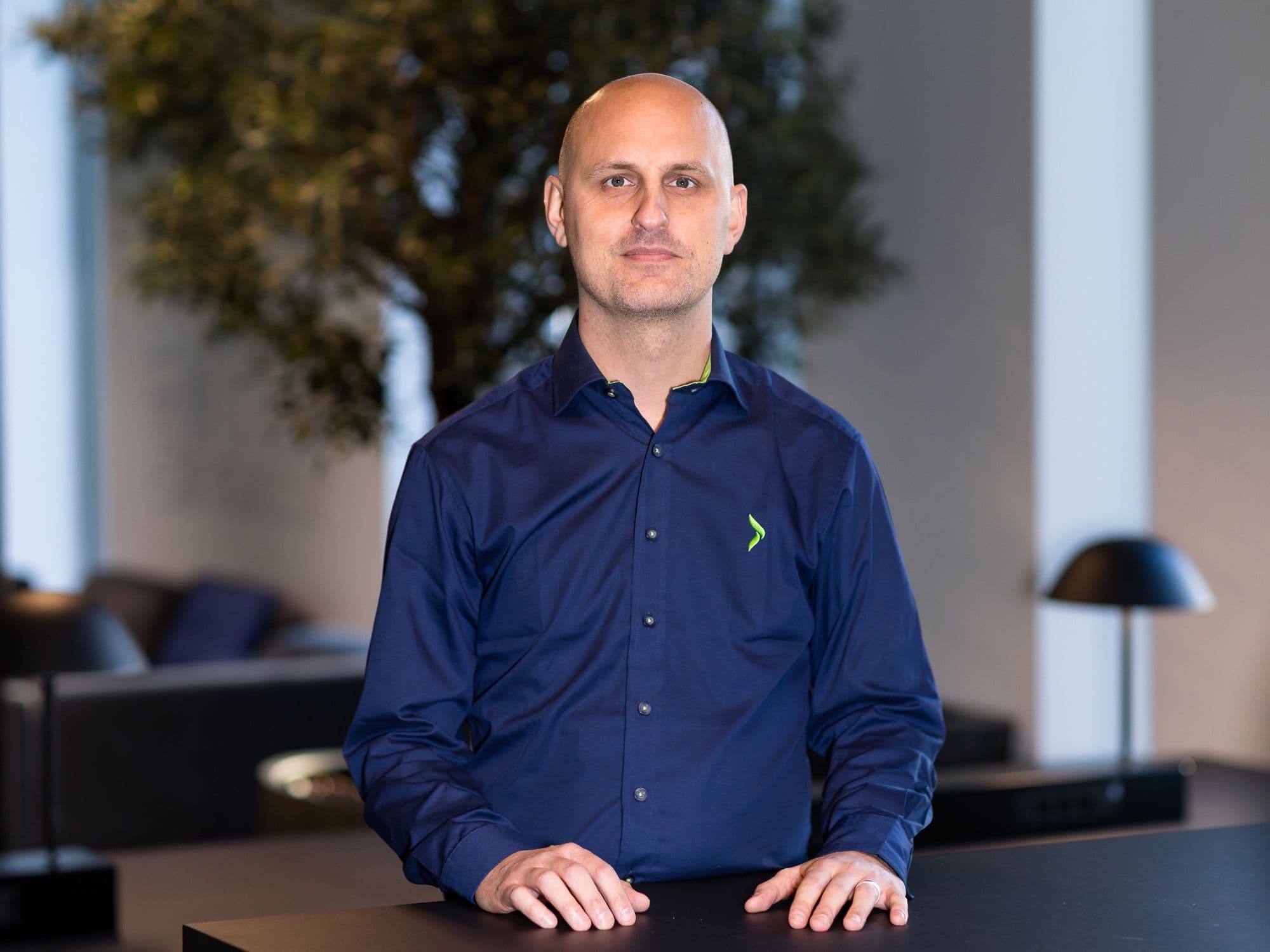 A bald man in a navy blue shirt with a green logo stands with his hands on a dark counter in a modern office.