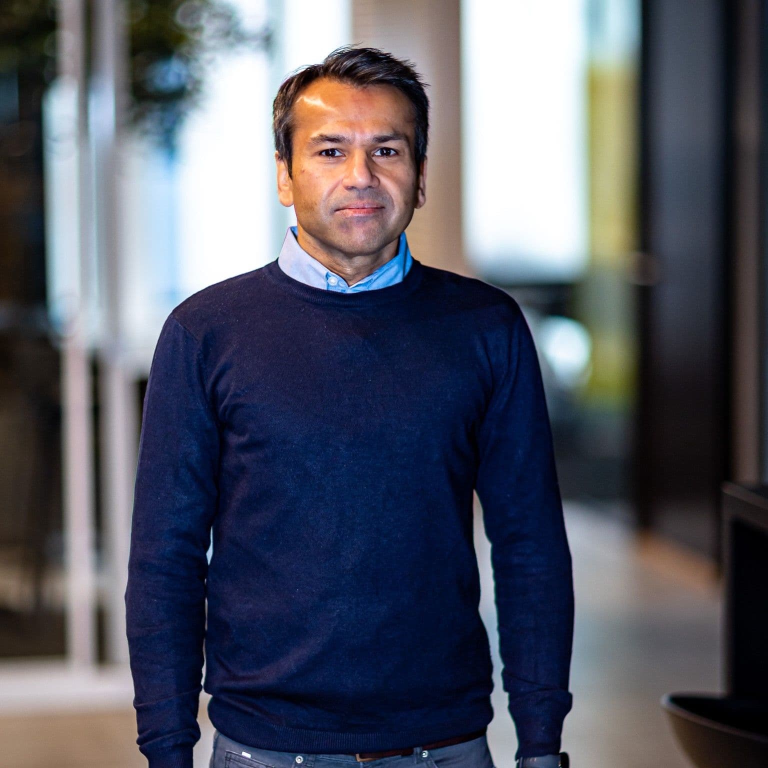 A man in a blue sweater and shirt stands looking at the camera.