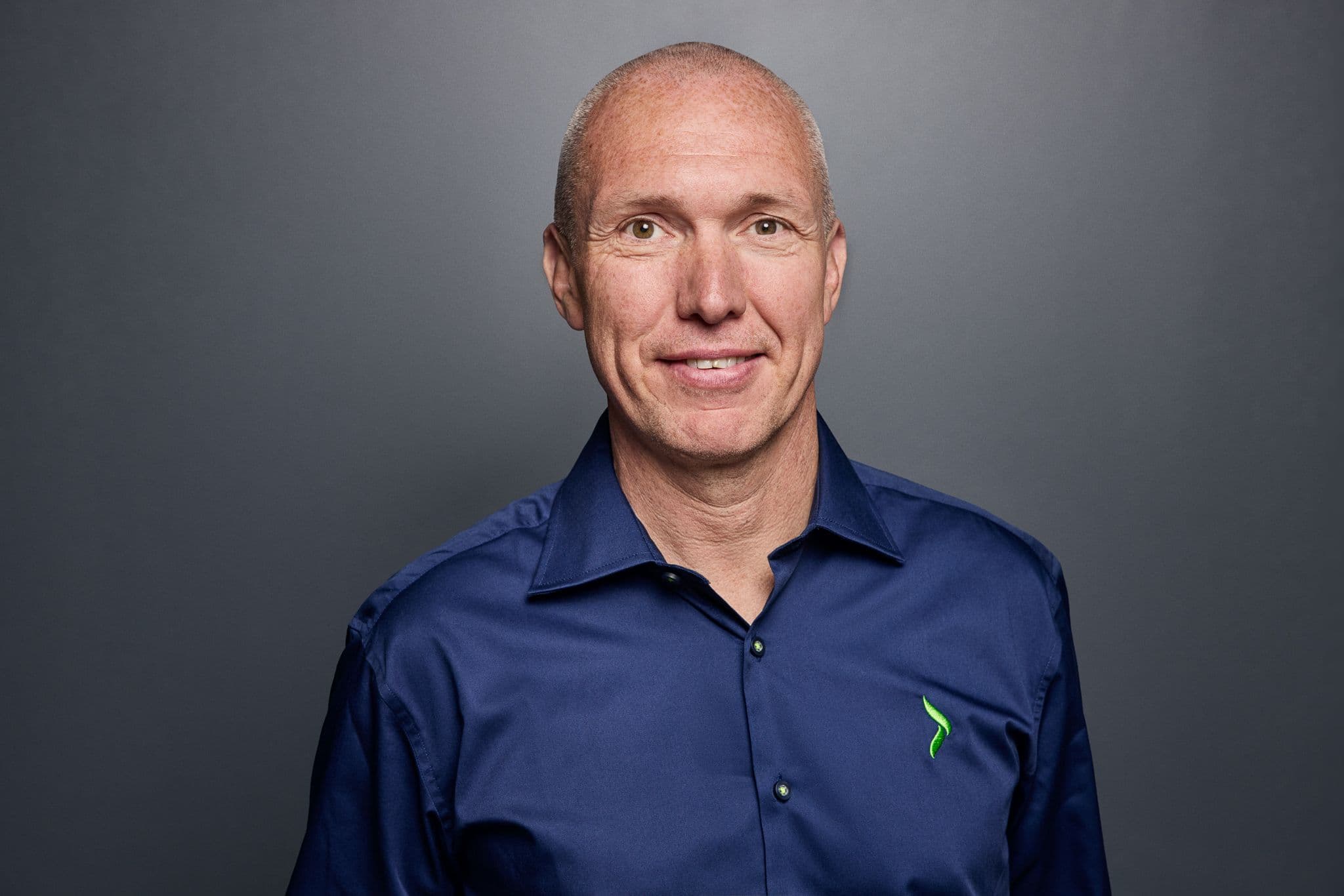 A smiling man in a blue collared shirt with a green logo.