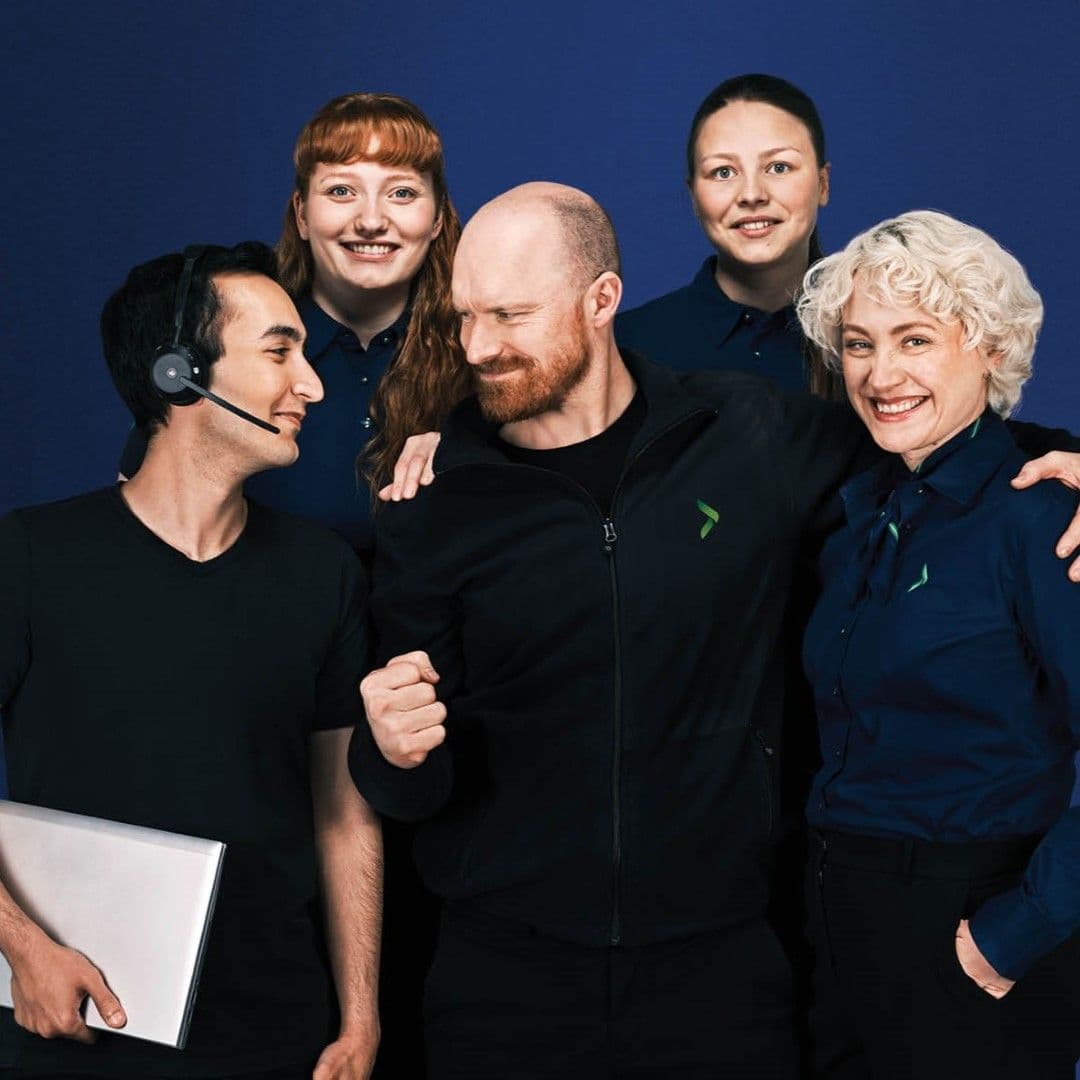 A smiling team of five people, two men and three women, wearing branded attire against a blue background. One man wears a headset and holds a laptop.