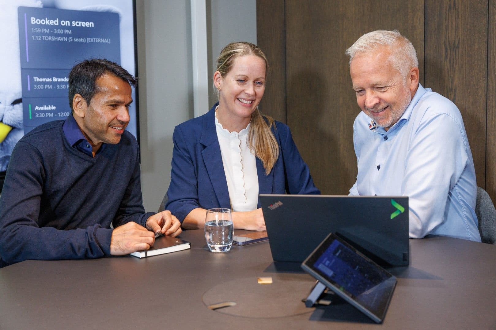 Three colleagues, two men and one woman, smiling and looking at a laptop in a meeting room.