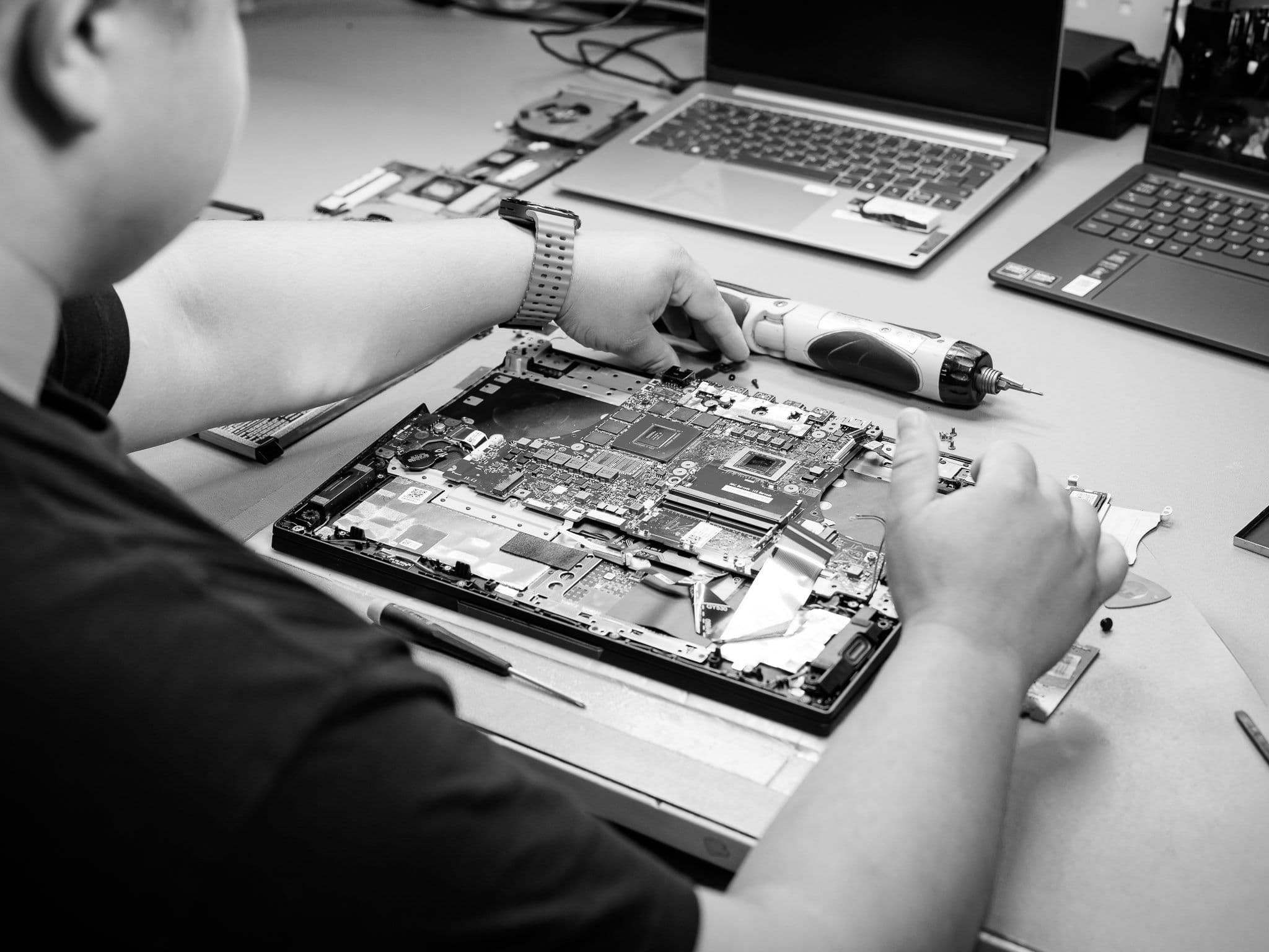A person disassembling a laptop, exposing its internal motherboard, with two other laptops in the background.